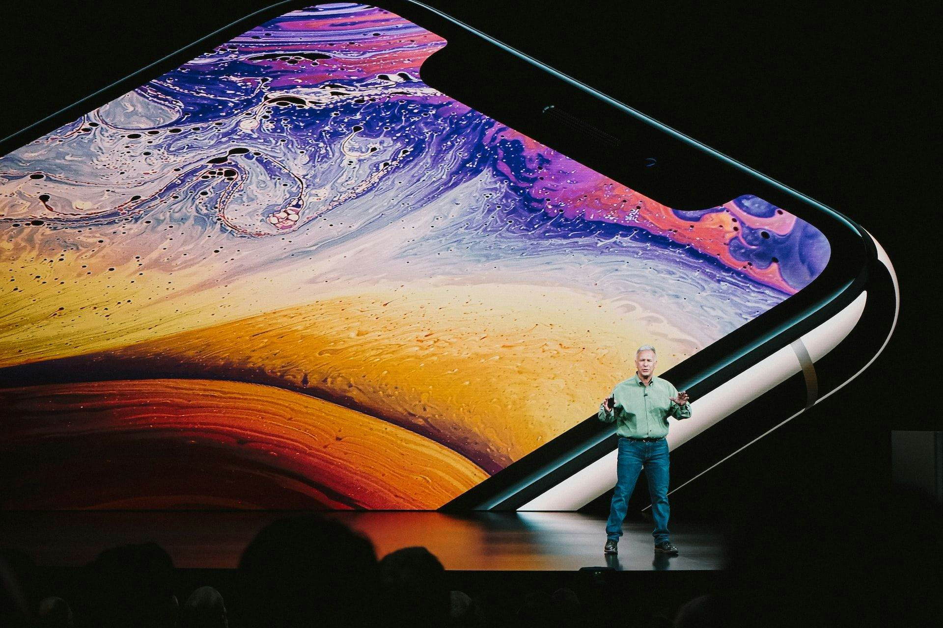 man speaking on stage with image of phone behind him